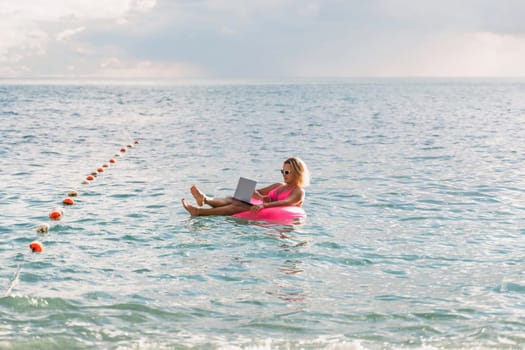 Woman works on laptop in sea. Freelancer, blond woman in sunglases floating on an inflatable big pink donut with a laptop in the sea at sunset. People summer vacation rest lifestyle concept