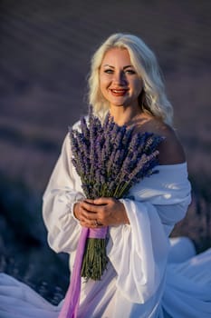 Blonde woman poses in lavender field at sunset. Happy woman in white dress holds lavender bouquet. Aromatherapy concept, lavender oil, photo session in lavender.