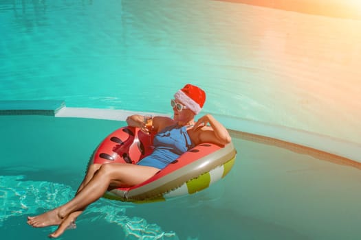 A happy woman in a blue bikini, a red and white Santa hat and sunglasses poses in the pool in an inflatable circle with a watermelon pattern, holding a glass of champagne in her hands. Christmas holidays concept