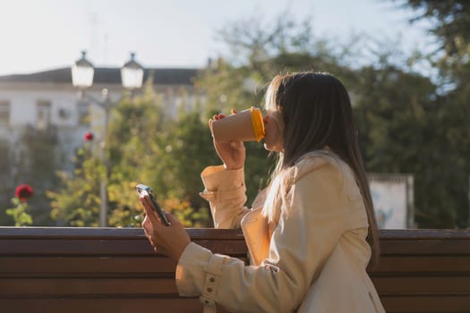 Woman drinks from cup on wooden bench. She is wearing a white shirt enjoying her beverage. The bench is located in a park setting, with trees in the background