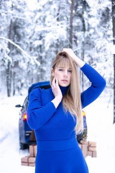 Woman in winter snowy forest in blue dress next to blue car decorated with Christmas decor. Christmas and winter holidays concept