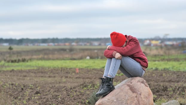 A sad teenage girl sits on a rock in a field and cries
