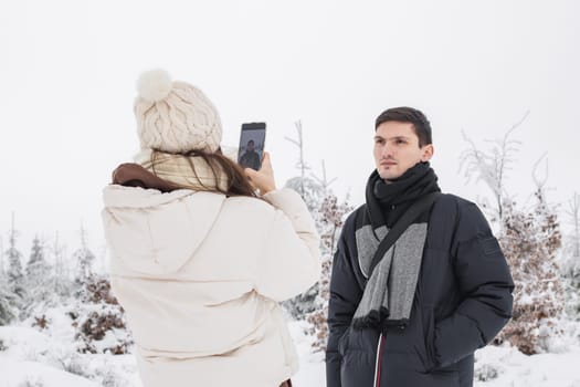 A young girl in winter clothes takes a picture on a smartphone of her handsome caucasian brunette boyfriend in a blue down jacket standing in a winter forest reserve in belgium, close-up side view. Concept of winter holidays, using technology.