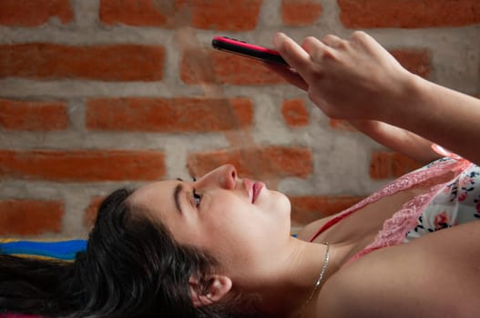 portrait of a girl from venezuela lying face up on her bed typing a text message with a black and pink cell phone. High quality photo