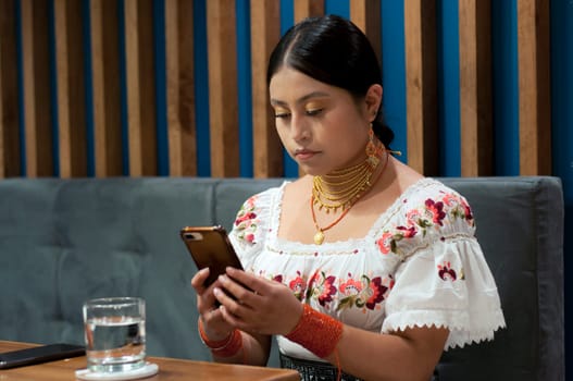 indigenous woman from otavalo, ecuador in traditional dress concentrating while writing a text message with her cell phone in a restaurant. High quality photo