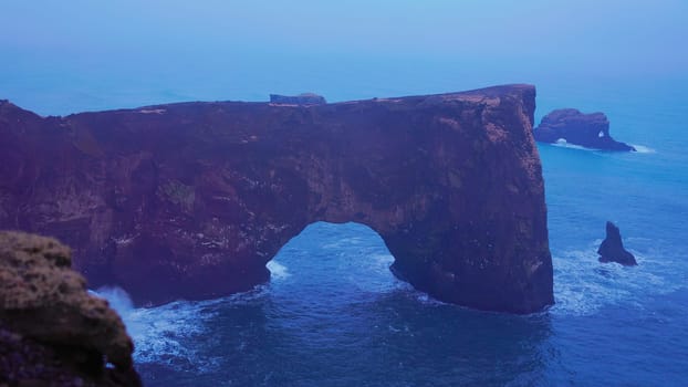 Stone coast arch on dyrholaey peninsula with natural foggy landscape and rock cliffs. Spectacular coastline gate in iceland, beautiful wild nature art with nordic design. Handheld shot.