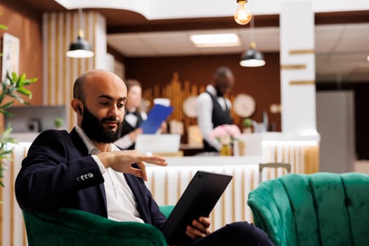 Middle eastern man on videocall using tablet, waiting to register in hotel lounge area. Employee travelling for business meetings attending online teleconference call with device.