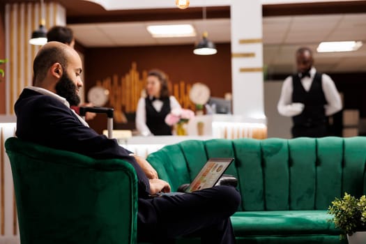 Middle eastern man using laptop in lobby, sitting in lounge area and preparing speech for international business meeting. Professional employee travelling for work, hotel reception.