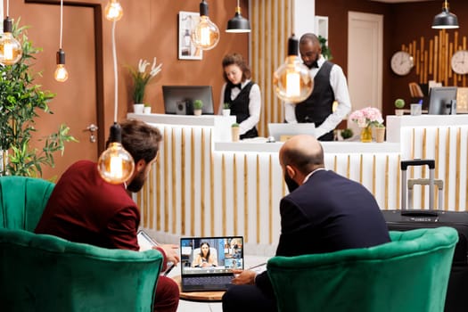 Business partners on videocall at hotel, talking to colleagues on online teleconference meeting. People in suits travelling for work and using videoconference call on laptop, lobby area.