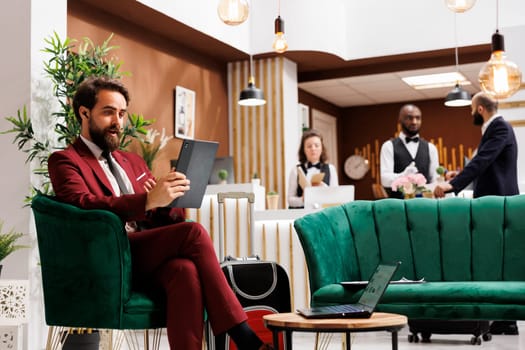 White collar worker talks on videocall, working abroad and travelling. Professional person in suit meeting online with partners to discuss new project ideas, using tablet in lobby at hotel.