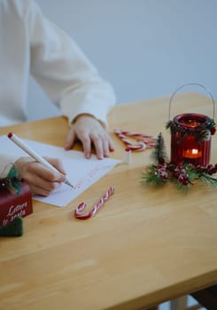 One unrecognizable Caucasian teenage girl writes a letter to dear Santa Claus with a red felt-tip pen, sitting at a wooden table with Christmas decorations, in the dining room on a winter day, side view from above, close-up.