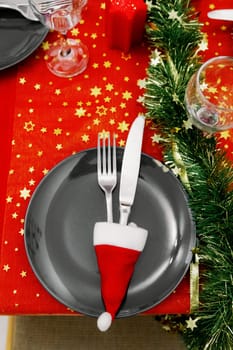 One gray plate with a knife and fork in a mini Santa Claus hat stand on a beautifully served holiday table with a red tablecloth and green tinsel on a winter evening in the kitchen, close-up top view with selective focus.