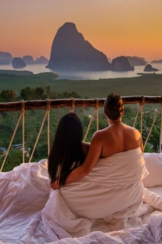 A couple in an outdoor bedroom watching the sunrise over Sametnangshe mountains in Phangnga Bay with mangrove forest in the Andaman Sea, men and woman in an outdoor bed at Phangnga, Thailand