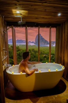 A man in a bathtub looking at the sunset over Sametnangshe viewpoint mountains in Phangnga Bay with a mangrove forest in the Andaman Sea in Phangnga, Thailand