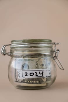 Saving Money In Glass Jar filled with Dollars banknotes. 2024 year transcription in front of jar. Managing personal finances extra income for future insecurity. Beige background