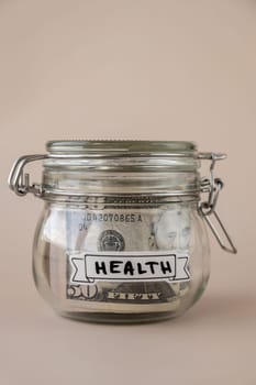 Saving Money In Glass Jar filled with Dollars banknotes. HEALTH transcription in front of jar. Managing personal finances extra income for future insecurity. Beige background