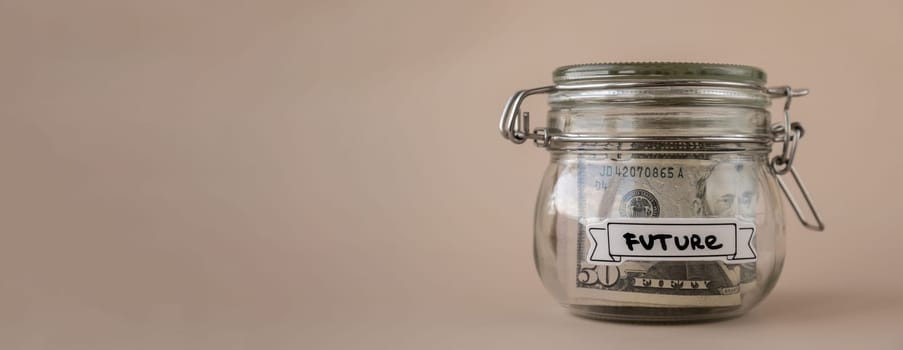 Saving Money In Glass Jar filled with Dollars banknotes. FUTURE transcription in front of jar. Managing personal finances extra income for future insecurity. Beige background