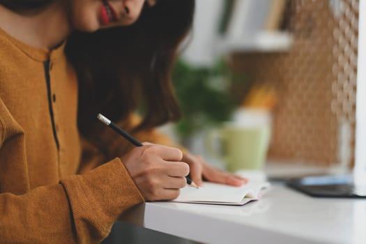 Cropped shot of young woman in yellow sweater writing in journal at her work desk.