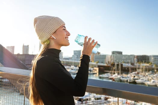 Happy young sporty woman taking a break from running drinking water outdoors during winter cold weather season. Copy space. Active lifestyle concept.