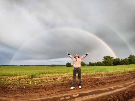 An adult girl in a field with stormy sky with clouds and grey rainbow. A woman having fun outdoors on rural and rustic nature near car. The Grey Rainbow and the Ban on LGBT symbols in Russia