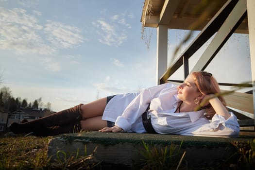 Beautiful girl with red hair in white shirt in open wooden pavillion in village or small town. Young slender woman and sky with clouds on background on autumn, spring or summer day