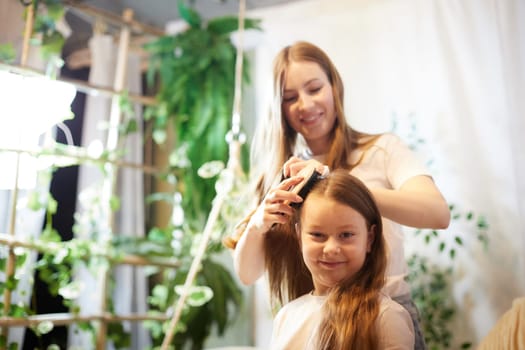 Young loving mom making ponytail to little schoolgirl daughter in living room. Mother helping child girl with a hairstyle