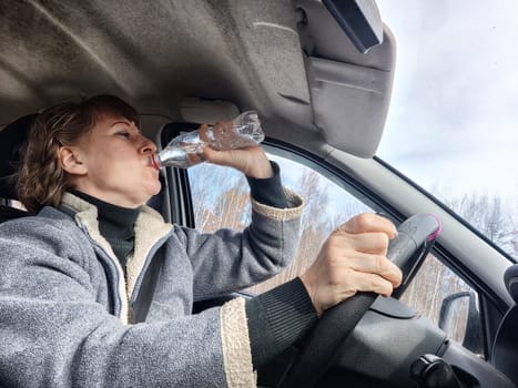 Portrait of female driver in solo journey. Adult mature woman holding steering wheel and bottle of water. Drinking while driving for to stay awake. Lady girl drinks alcohol vodka or gin while driving