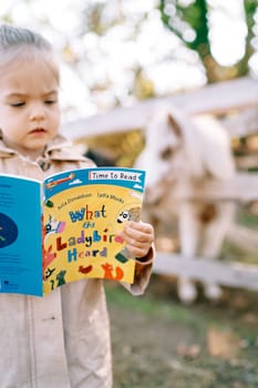 Little girl enthusiastically reads a picture book while standing in the ranch. High quality photo