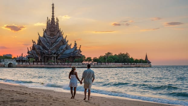 A diverse multiethnic couple of men and women visit The Sanctuary of Truth wooden temple in Pattaya Thailand at sunset, couple watching sunset on the beach in Pattaya with a wooden temple