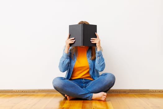 A young woman covers her face with a book as she sits on the floor against the wall.