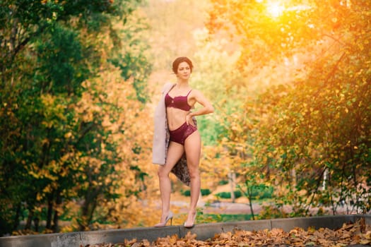 Young beautiful dark-haired woman with a slender figure posing in lingerie and a classic coat in an autumn park