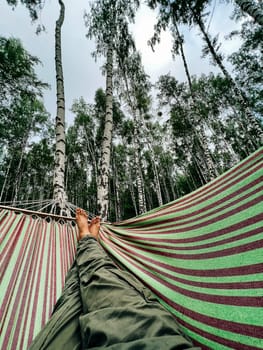 Legs of man, swinging on hammock at summer in birch forest. Enjoying, guy dreaming, resting under tree. Happy lifestyle. High quality