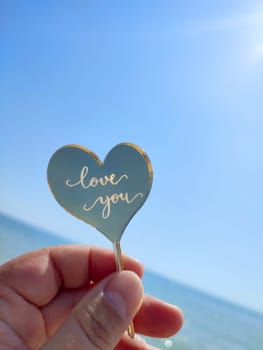 Person holding in fingers hand stick in shape heart and words Love you on background sea and sea waves, blue sky in seashore on sunny summer day close-up. Concept love romance amour St Valentines Day