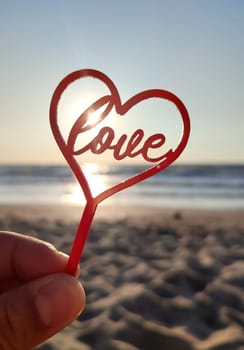 Person holding in fingers hand stick in shape red heart and word Love on background sea and sea waves, blue sky in seashore on sunny summer day close-up. Concept love romance amour St Valentines Day