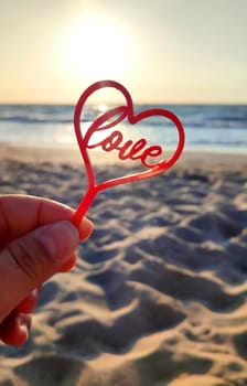 Person holding in fingers hand stick in shape red heart and word Love on background sea and sea waves, blue sky in seashore on sunny summer day close-up. Concept love romance amour St Valentines Day
