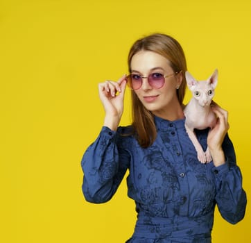 Happy blond woman Egyptian cat owner with pet on shoulder isolated on yellow background. Exotic cat ownership concept. High quality photo