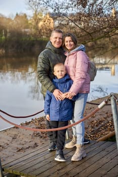 Young parents and their little son standing on the pier near the lake or river shore, at sunset on autumn day.