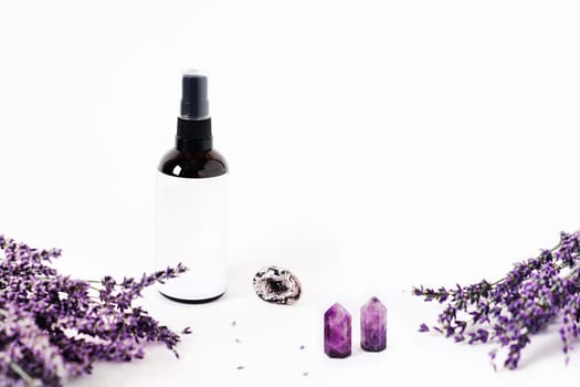 Glass spray bottle on white background surrounded by lavender bouquets and the crystal of quartz. Energy of stones for your home.