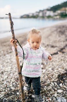 Little girl stands on a pebble beach with a big stick. High quality photo