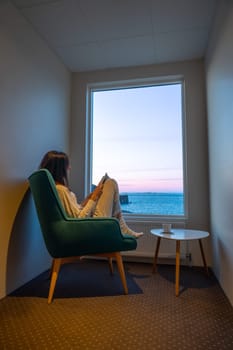 Woman in pajamas reading in a window facing the sea at sunset