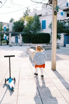 Little girl walks in the courtyard of the house near a three-wheeled scooter. Back view. High quality photo