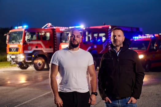 Group of firefighters, dressed in civilian clothing, stand in front of fire trucks during the night, showcasing a moment of camaraderie and unity among the team as they reflect on their duties and the challenges faced during their firefighting service.