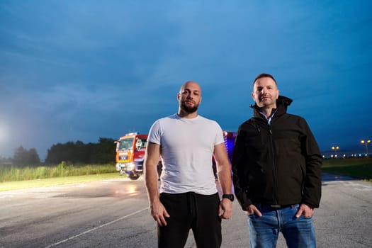 Group of firefighters, dressed in civilian clothing, stand in front of fire trucks during the night, showcasing a moment of camaraderie and unity among the team as they reflect on their duties and the challenges faced during their firefighting service.