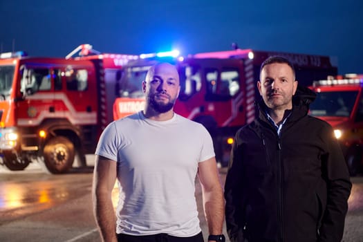 Group of firefighters, dressed in civilian clothing, stand in front of fire trucks during the night, showcasing a moment of camaraderie and unity among the team as they reflect on their duties and the challenges faced during their firefighting service.