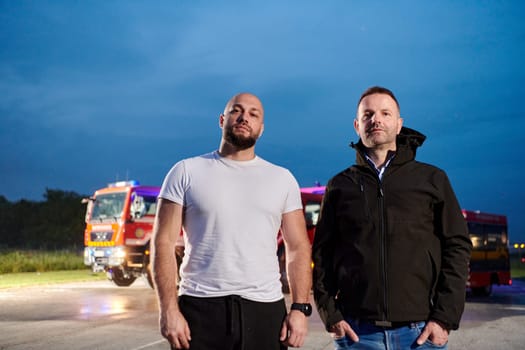 Group of firefighters, dressed in civilian clothing, stand in front of fire trucks during the night, showcasing a moment of camaraderie and unity among the team as they reflect on their duties and the challenges faced during their firefighting service.