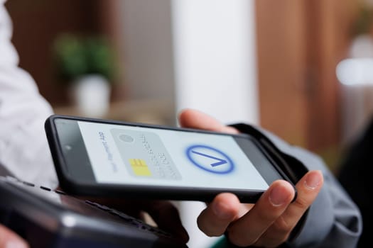 Close-up shot of tourist making contactless payment via mobile phone using NFC technology. Detailed view of pair of hands grasping pos terminal and smartphone, paying winter holiday reservation.