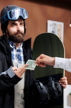Detailed view of male snowboarder tipping employee with cash in ski hotel reception. Caucasian man in wintersport clothing giving gratuity to concierge at winter mountain resort.