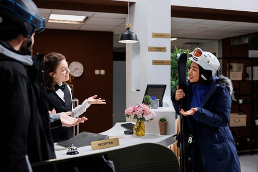 Friendly receptionist assisting female tourist with reservation and information on best skiing slopes at winter mountain resort. Young travelers carrying wintersports equipment talking with concierge.