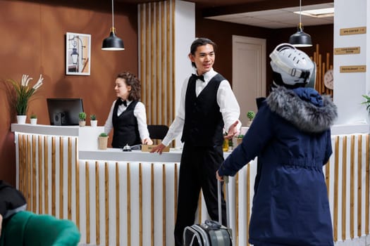 Asian male concierge assists woman in winter clothing with luggage at hotel reception. Female traveler approaches front desk of ski mountain resort with suitcase and receives assistance from employee.