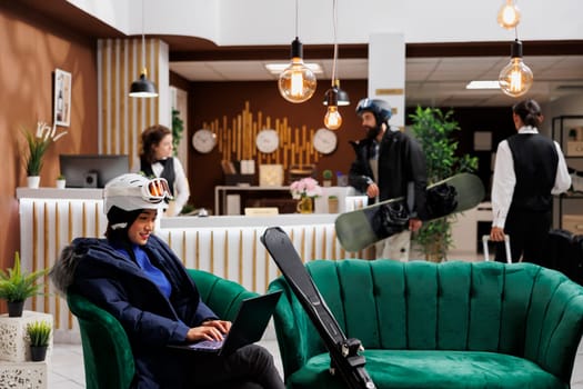 Woman wearing ski goggles and winter gear sits in lounge area, engrossed in laptop, planning skiing adventure. At front desk, guest discusses booking with receptionist for winter travel at ski resort.